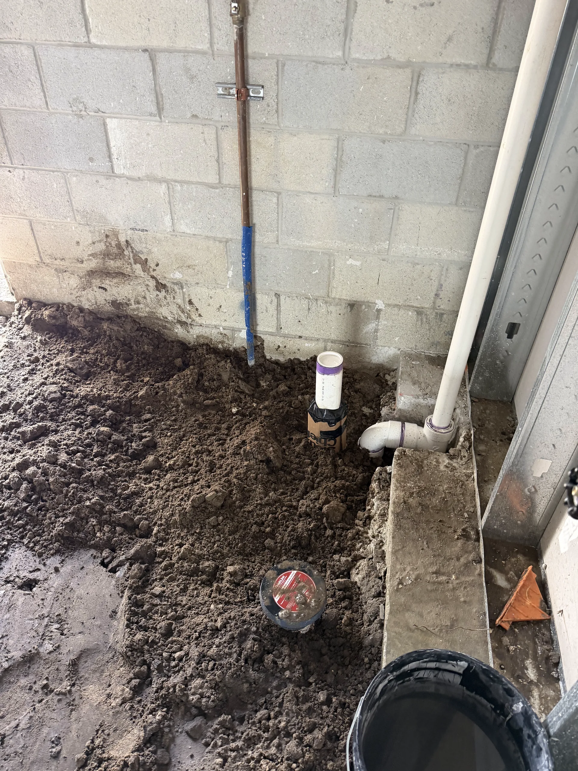 Hand-dug trench along a block wall with the plumbing stub and cleanout set into the fill