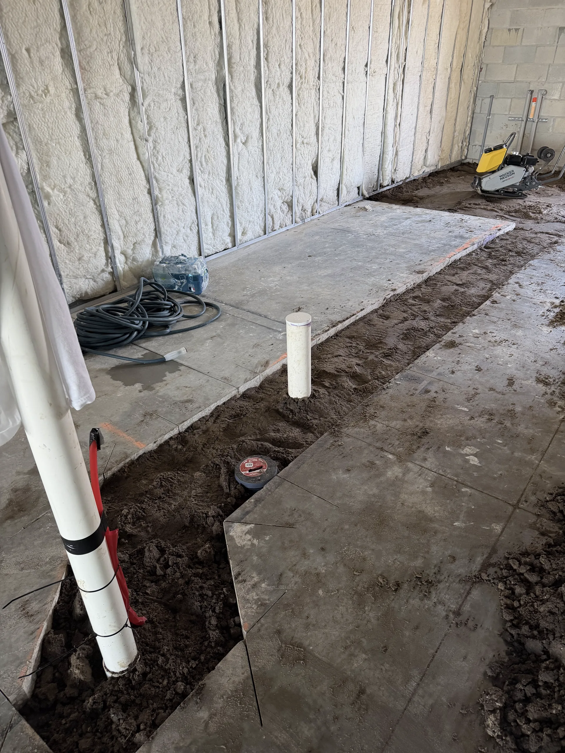 Concrete slab sections removed and a trench dug for plumbing rough-in, plate compactor on the fill